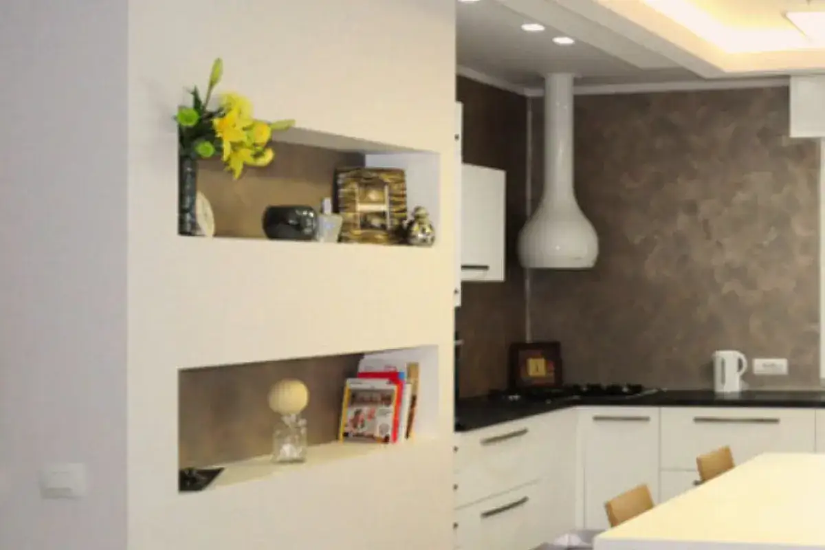 Parete divisoria in cartongesso con nicchie che espongono decorazioni e libri. La cucina moderna &egrave; visibile sullo sfondo.
