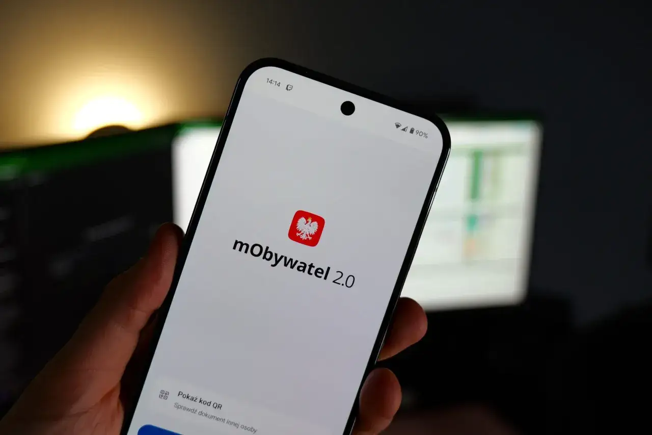 Dłoń trzyma smartfon z aplikacją mObywatel 2.0. Na ekranie widać logo aplikacji i opcję