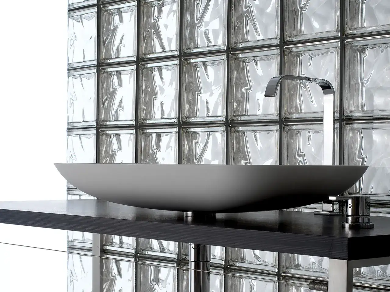 Lavabo moderno su mobile scuro, con rubinetto cromato e un suggestivo muro di vetrocemento retrostante che riflette la luce.