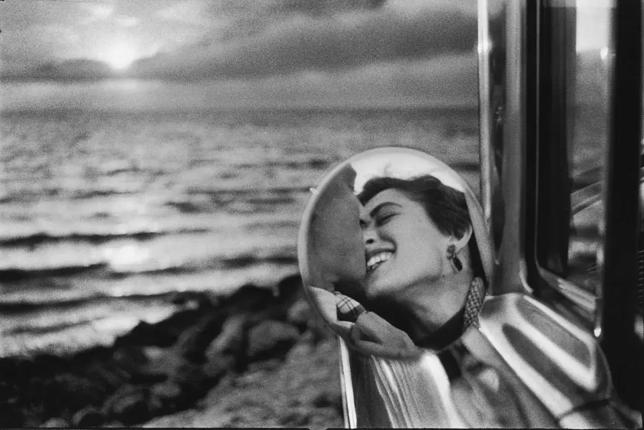 Un baiser volé, capturé par le plus grand photographe du monde. Une femme rit, son visage reflété dans un rétroviseur, la mer en arrière-plan.