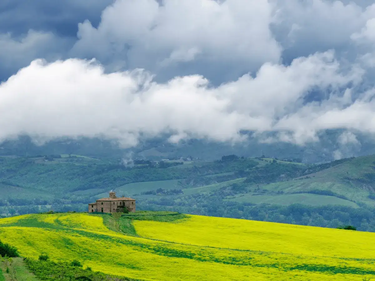 Un casale solitario su una collina fiorita di giallo, immerso nel paesaggio del cammino dei borghi silenti, sotto un cielo nuvoloso.