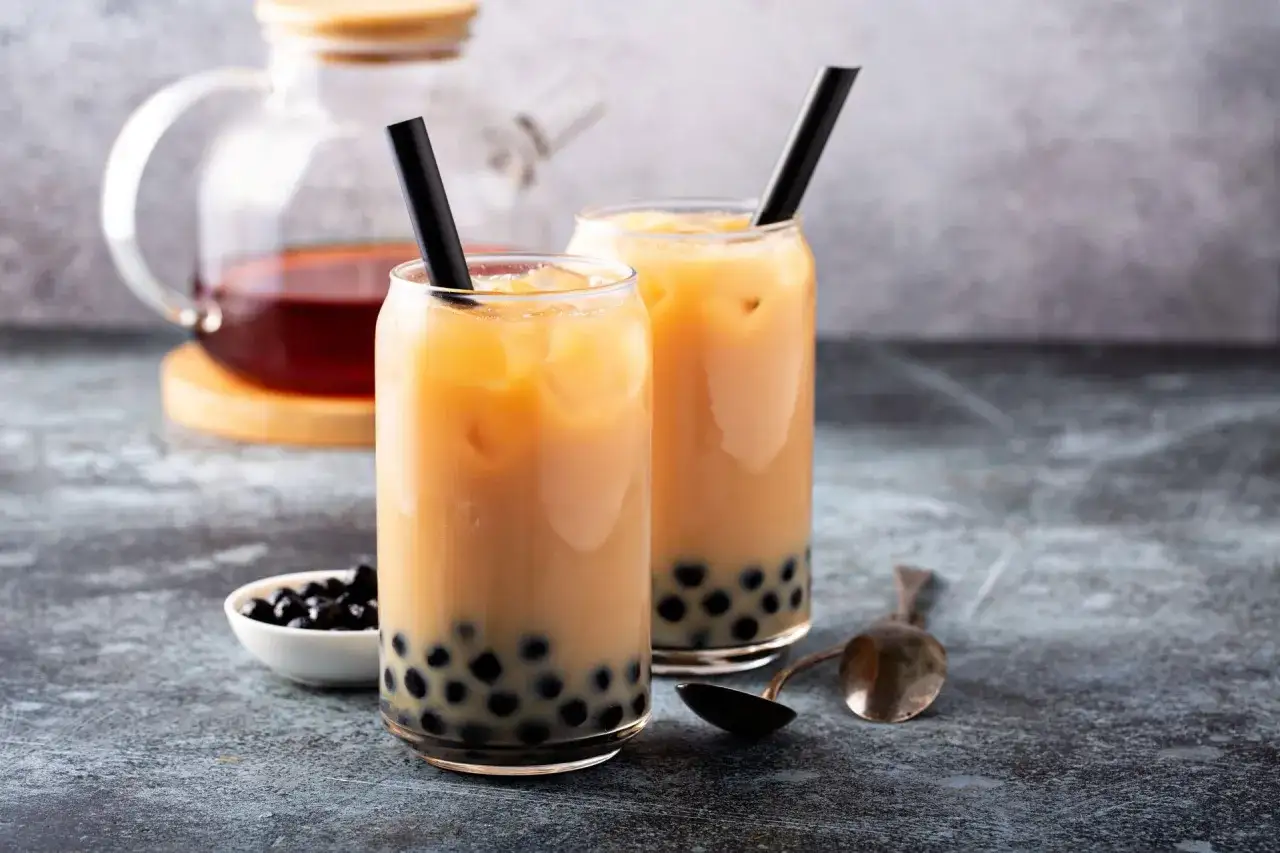 Dwa szklanki orzeźwiającej bubble tea z czarnymi tapiokowymi kuleczkami i czarnymi słomkami. Obok miseczka z kuleczkami i łyżeczka.