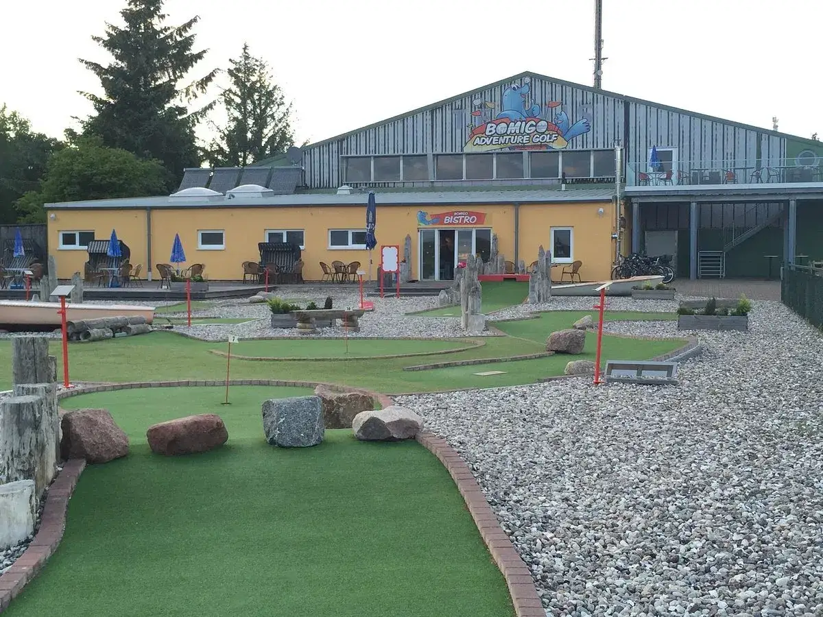 BOMIGO Adventure Golf Zingst: Eine Minigolfanlage mit Bistro, umgeben von Kies und grünen Kunstrasenbahnen.