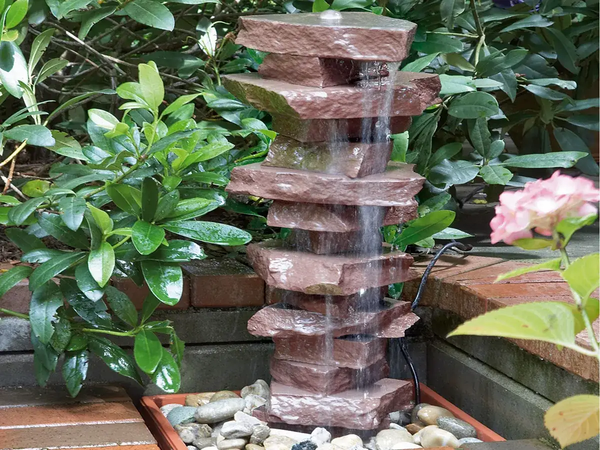 Ein gestapelter Stein-Gartenbrunnen, der zeigt, wie man einen Gartenbrunnen selber bauen kann. Wasser flie&szlig;t &uuml;ber die Steine in ein Kiesbett.