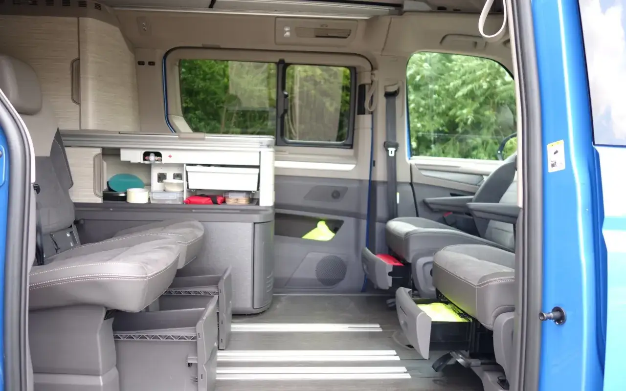 Wnętrze kampera Volkswagen California. Widok na fotele, kuchnię i schowki. Idealny do podróży, a jego **volkswagen california cena** jest atrakcyjna.
