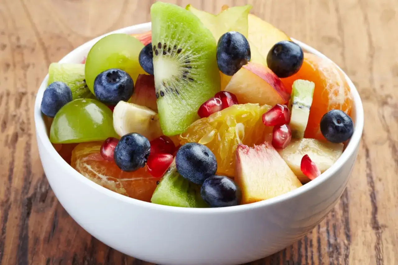 Kolorowa sałatka owocowa zimowa: kiwi, pomarańcza, winogrona, borówki, granat, brzoskwinia i banan w białej misce.
