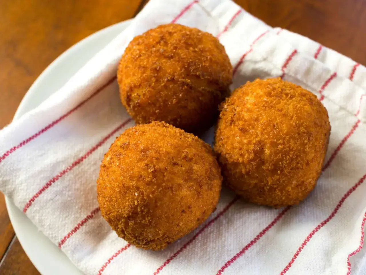 Trzy złociste, chrupiące włoskie kotlety z ryżu (arancini) na białej serwetce z czerwonymi paskami.