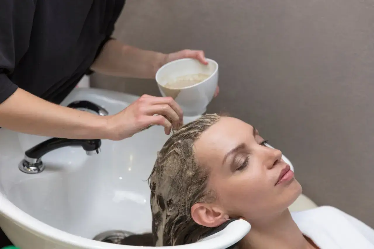 Una peluquera aplica un tratamiento capilar a una mujer en un lavacabezas. La pregunta de si es mejor teñirse con el pelo sucio o limpio es clave para el resultado.