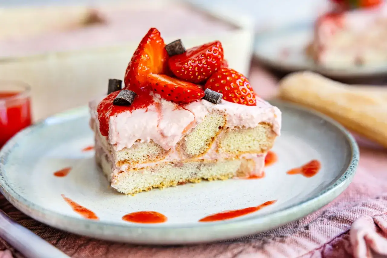 Ein Stück Erdbeer-Tiramisu ohne Ei, dekoriert mit frischen Erdbeeren und Schokostückchen, auf einem Teller mit Erdbeersauce.