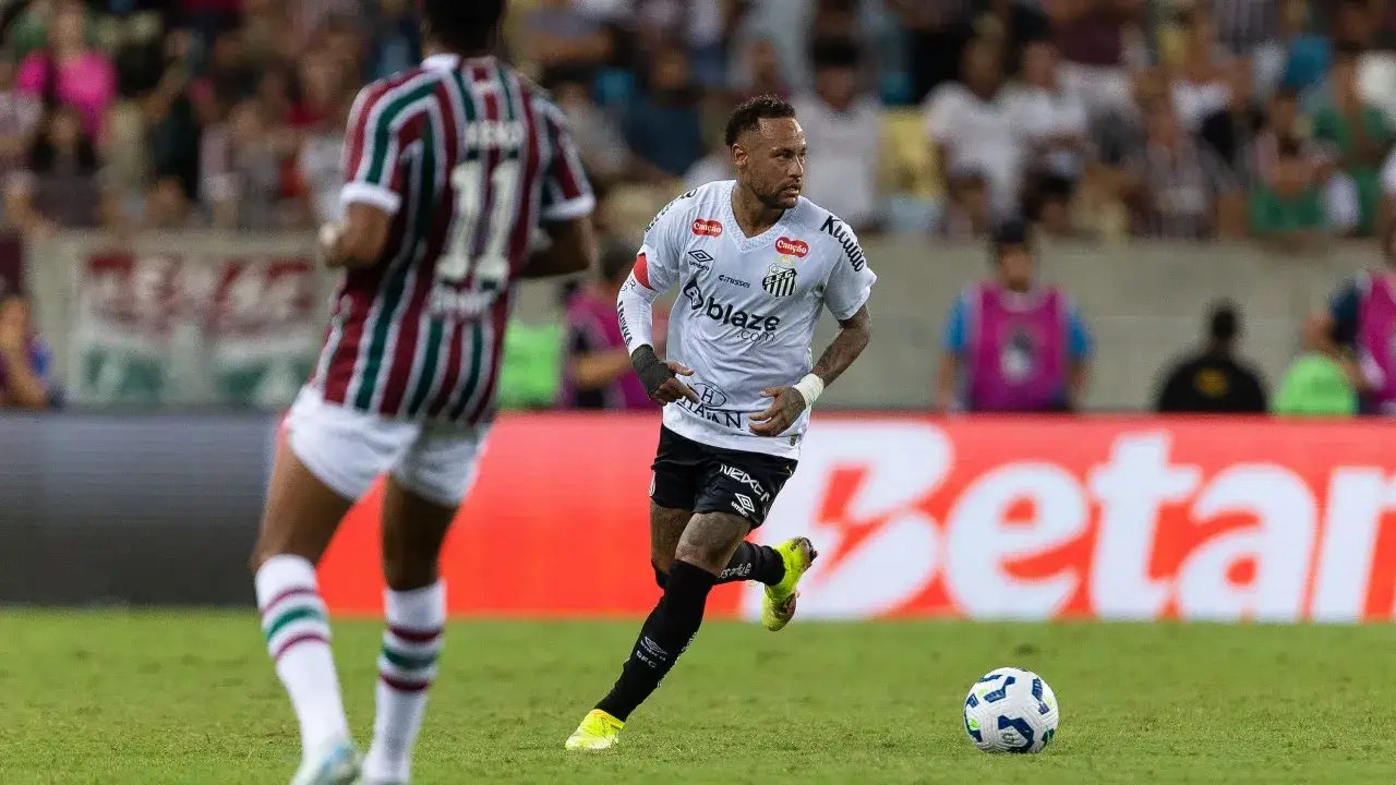Neymar w białej koszulce biegnie po boisku, piłka tuż obok. To właśnie tam gra Neymar, w akcji meczowej.