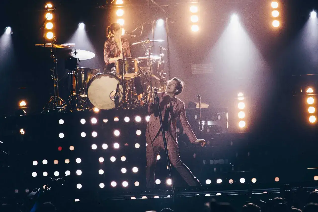 Harry Styles uppträder i Stockholm, med en trummis i bakgrunden. Scenen är upplyst av många lampor.