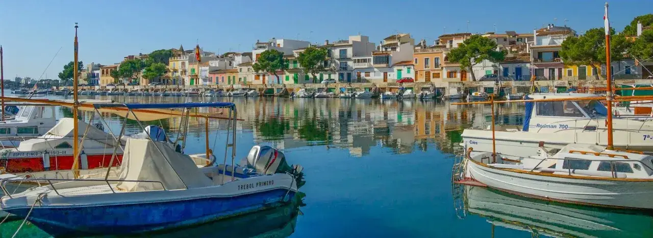 Boote im Hafen von Portocolom, Mallorca, spiegeln sich im ruhigen Wasser vor bunten H&auml;usern.