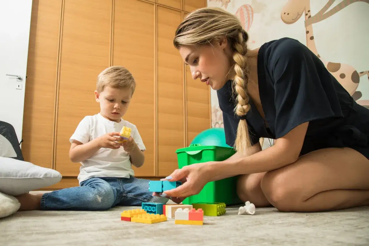 Mutter und Sohn bauen mit bunten Kl&ouml;tzen. Ein kleiner Junge h&auml;lt einen gelben Klotz, w&auml;hrend die Mutter ihm einen blauen gibt. Der **Trennungsschmerz Kindergarten** ist hier nicht zu sp&uuml;ren, nur Spiel und Spa&szlig;.