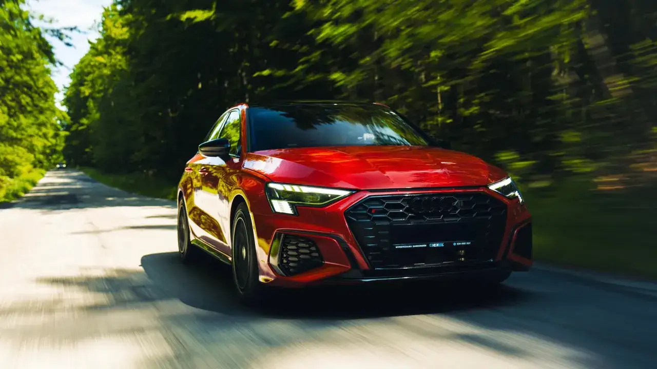 Czerwony audi sedan pędzi leśną drogą, rozmazując zieleń drzew.