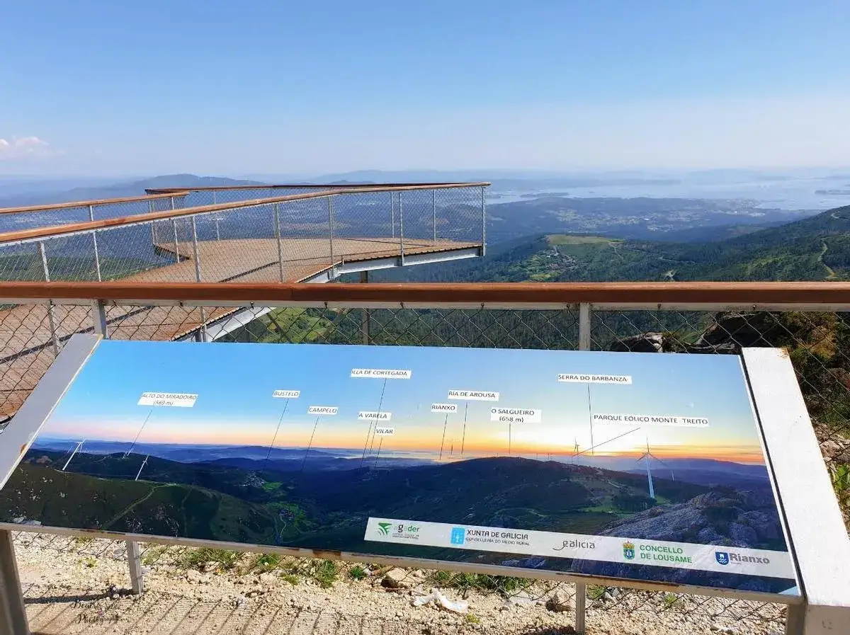 Mirador Monte Muralla: una pasarela de madera se adentra en un paisaje montañoso con vistas al mar. Un panel informativo muestra la panorámica.