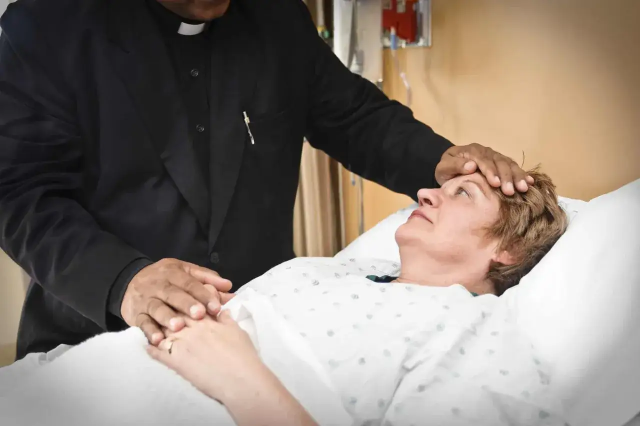 A priest performs the anointing of the sick, placing his hands on a woman lying in a hospital bed, offering comfort and spiritual solace.