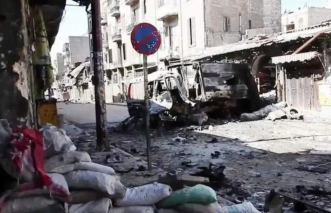 Spalony pojazd i zniszczone budynki na ulicy. Worek z piaskiem i czerwona tkanina na pierwszym planie. Czy w Syrii jest bezpiecznie?