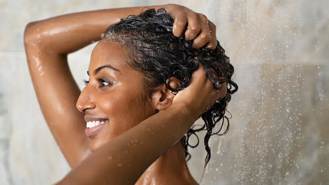 Une femme souriante se lave les cheveux sous la douche. Elle a les cheveux foncés et ondulés. Elle semble apprécier ce moment, peut-être une fois toutes les deux semaines.