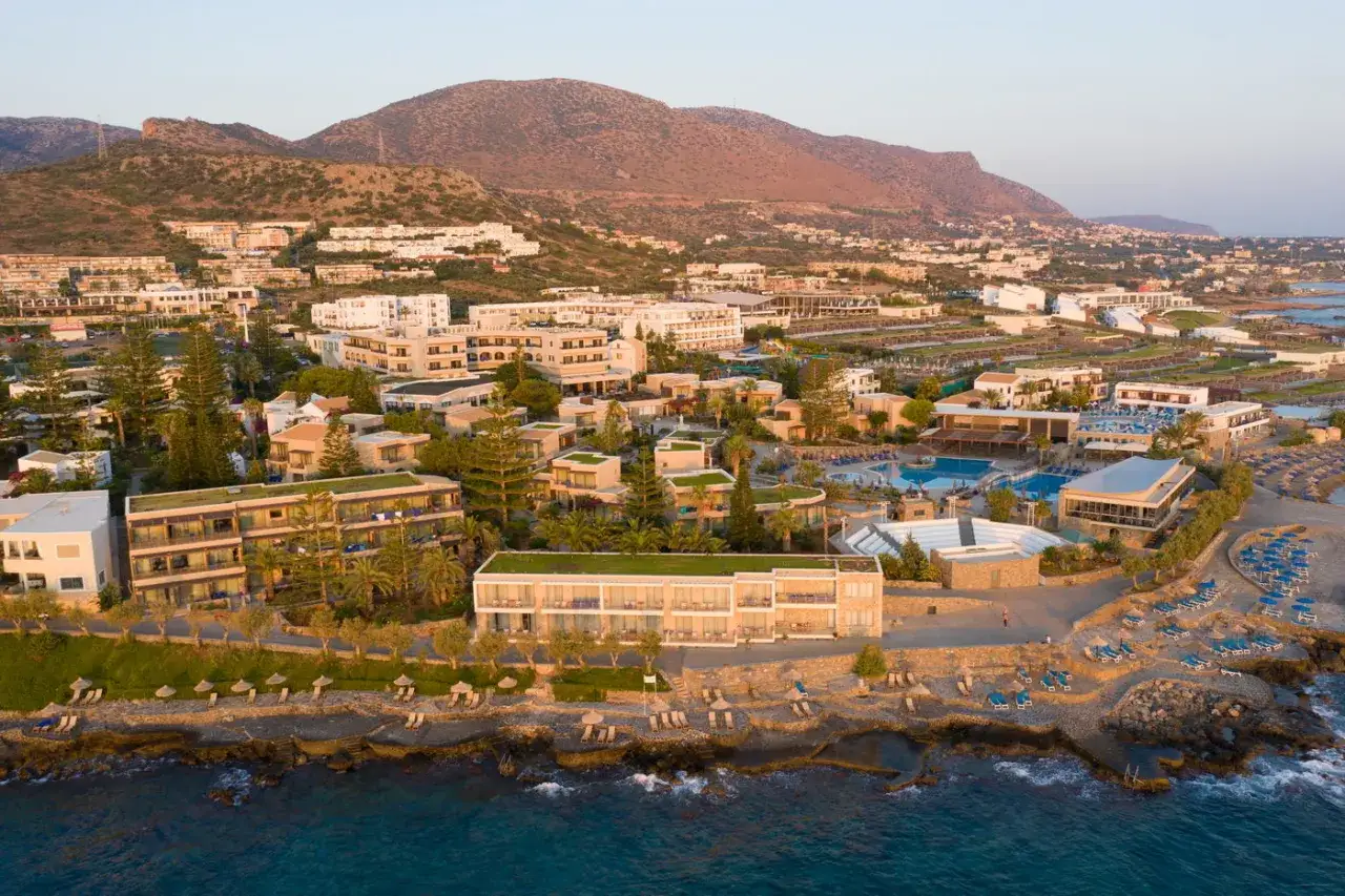Luftaufnahme eines Resorts am Nana Beach Kreta. Mehrere Gebäude, Pools und Liegestühle sind zu sehen, mit Bergen im Hintergrund.