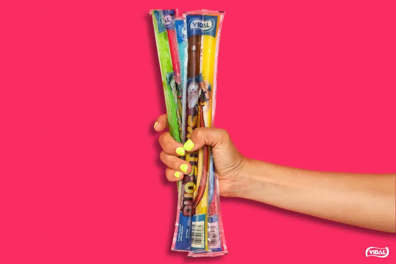 Mano con uñas amarillas sosteniendo varios helados flash de Vidal de diferentes sabores, sobre fondo rosa vibrante.
