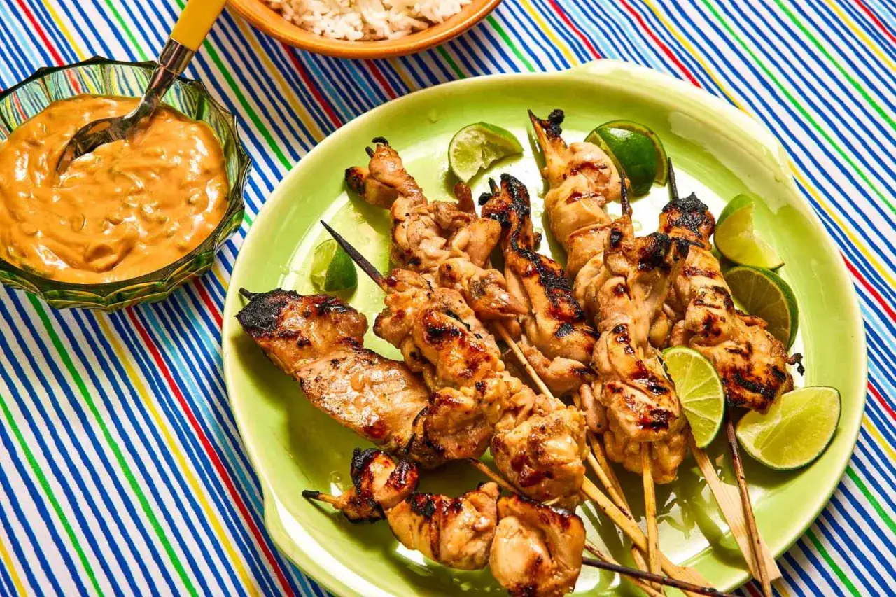 Saftige Hähnchen-Satay-Spieße mit Erdnusssauce und Limettenspalten auf einem bunten Tischläufer.