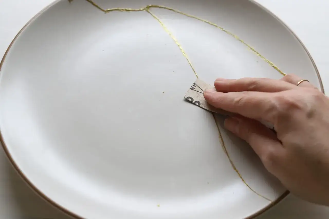 Dłoń z papierem ściernym delikatnie wygładza złote linie na pękniętej porcelanie, tworząc unikalne dzieło sztuki. Klej do porcelany pozwala na odnowę.