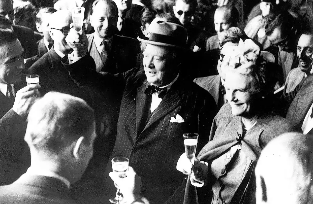 Churchill, chapeau sur la tête, lève son verre. Une foule joyeuse l'entoure, célébrant avec du **churchill alcool**.