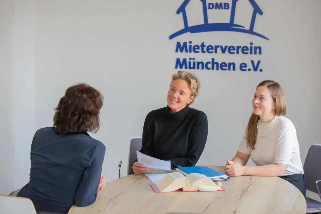Drei Personen besprechen Dokumente am Tisch. Im Hintergrund das Logo des Mietervereins München e.V. – die Frage welcher Mieterverein ist der beste wird hier vielleicht geklärt.