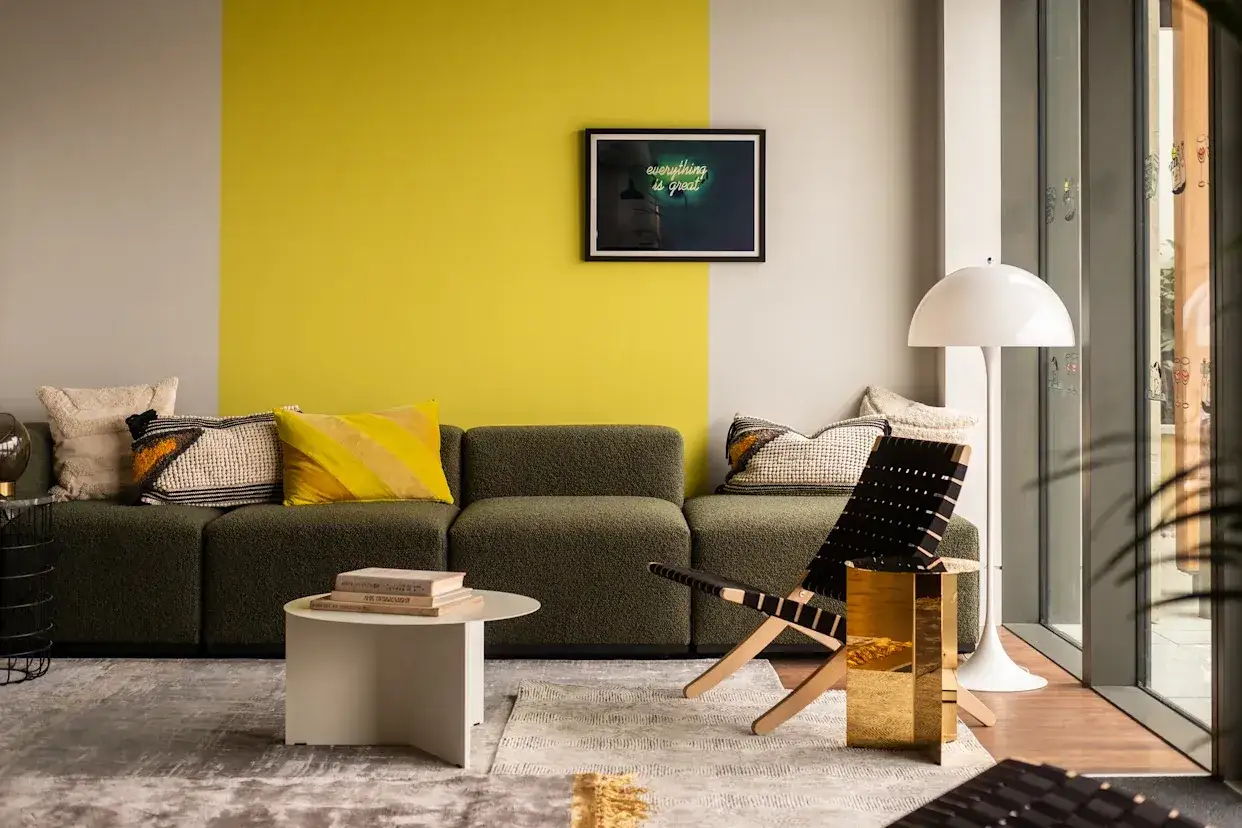 En modern soffgrupp med en gr&ouml;n soffa, kuddar i olika m&ouml;nster och f&auml;rger, ett vitt soffbord och en golvlampa. V&auml;ggen &auml;r m&aring;lad i tv&aring; f&auml;rger, ljusgr&aring;tt och knallgult, vilket ger en k&auml;nsla av att m&aring;la kanter utan tejp.