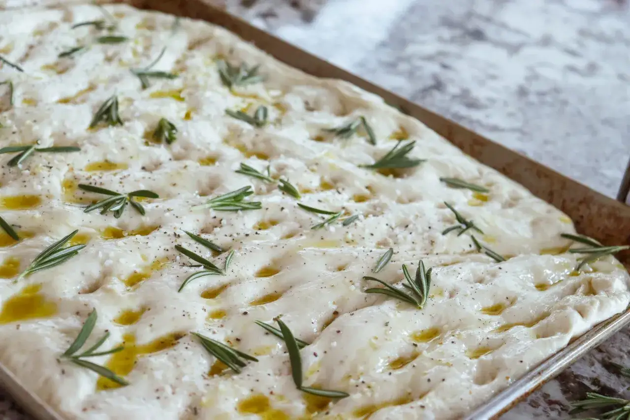 Focaccia rosmarin p&aring; en pl&aring;t, redo att bakas. Olivolja, flingsalt och peppar toppar det fluffiga br&ouml;det.