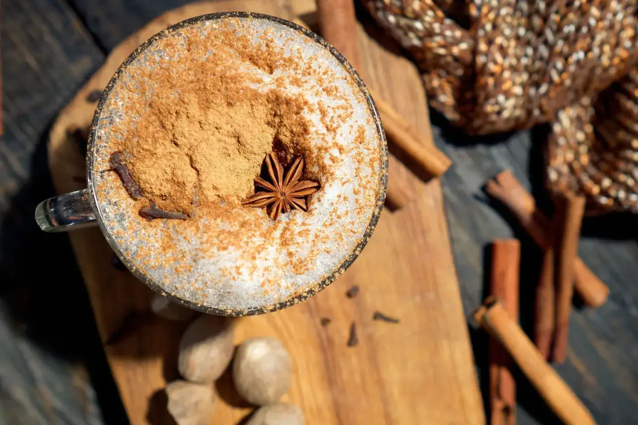 Ein Glas Chai Latte mit Zimt, Sternanis und Nelken. Die chai latte inhaltsstoffe sind hier sichtbar: Zimtstangen, Muskatnüsse und ein Gewebestück.