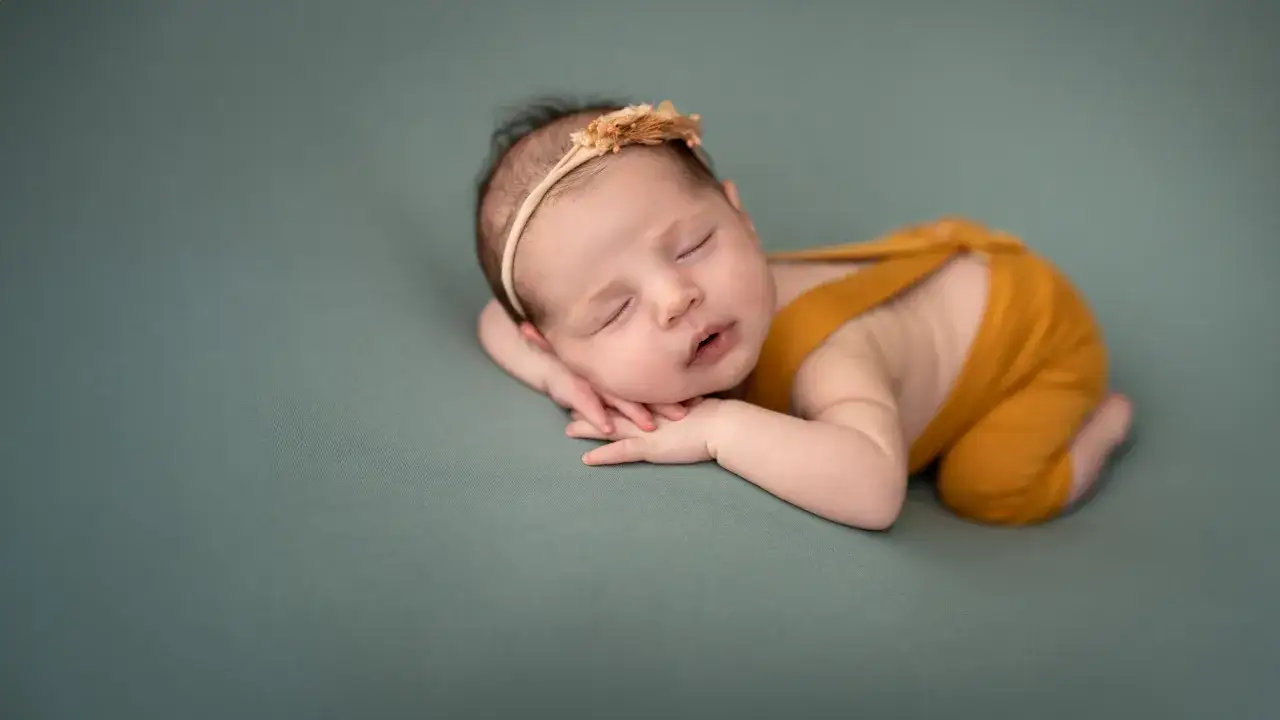 Dolce neonato scatti nel sonno, con una coroncina floreale e un pagliaccetto color senape, adagiato su un morbido sfondo verde salvia.
