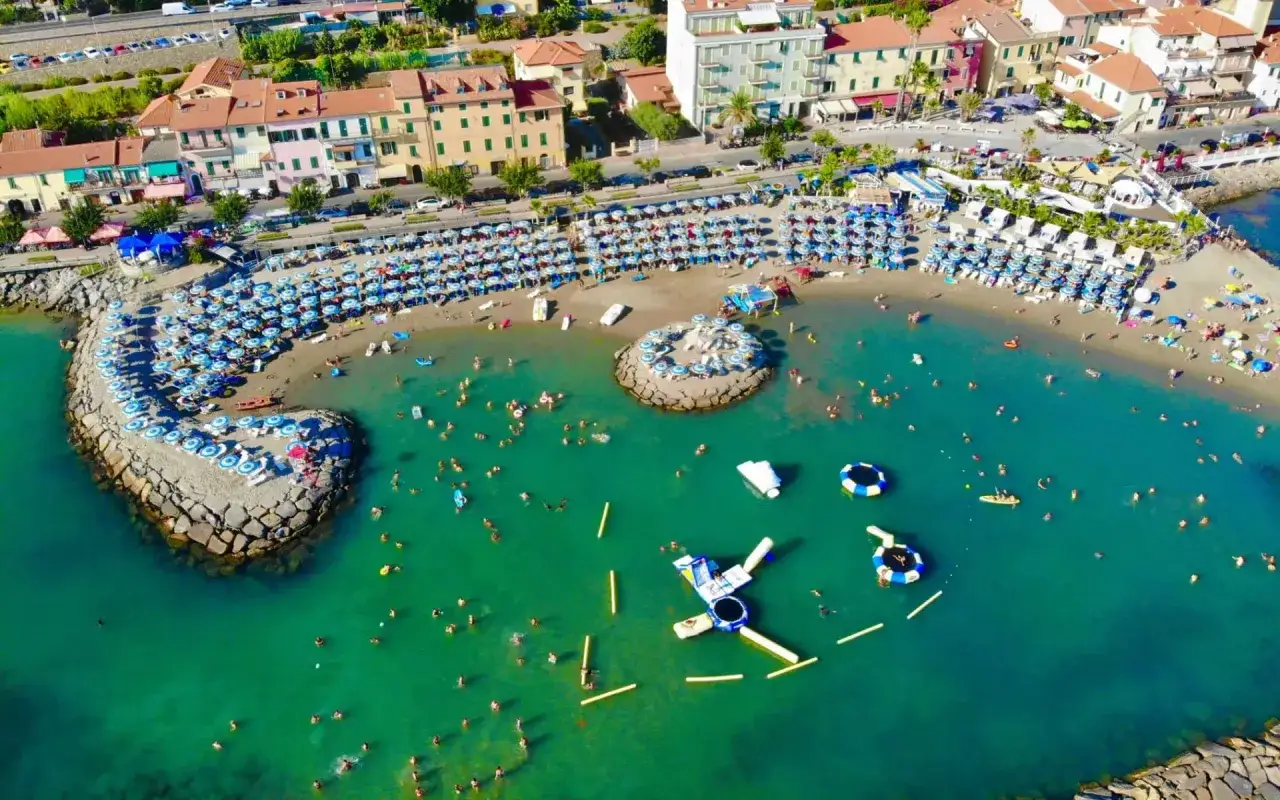 Plage anim&eacute;e &agrave; Arma di Taggia, Italie, avec des parasols bleus et blancs, des baigneurs dans la mer turquoise et des structures gonflables.