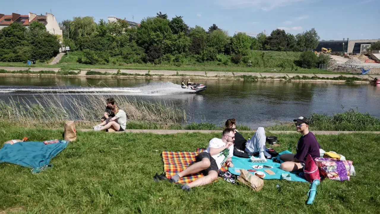 Ludzie odpoczywają nad rzeką, ciesząc się majówką. Na wodzie śmiga skuter wodny, a na trawie piknikują grupy znajomych.