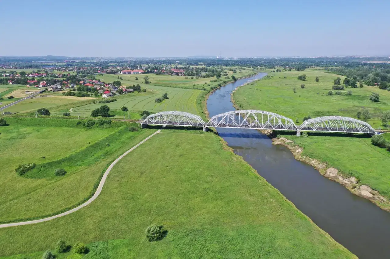 Malownicza wiślana trasa rowerowa Kraków-Niepołomice. Most kolejowy nad Wisłą, zielone łąki i wiejska zabudowa.