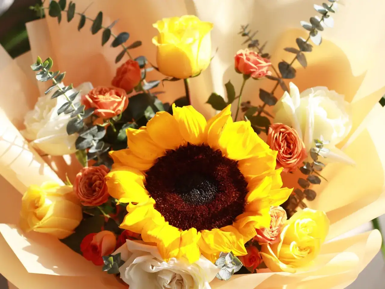 Un ramo de girasoles y rosas naranjas y amarillas, envuelto en papel color melocot&oacute;n. El girasol, s&iacute;mbolo de felicidad, ilumina este arreglo floral.
