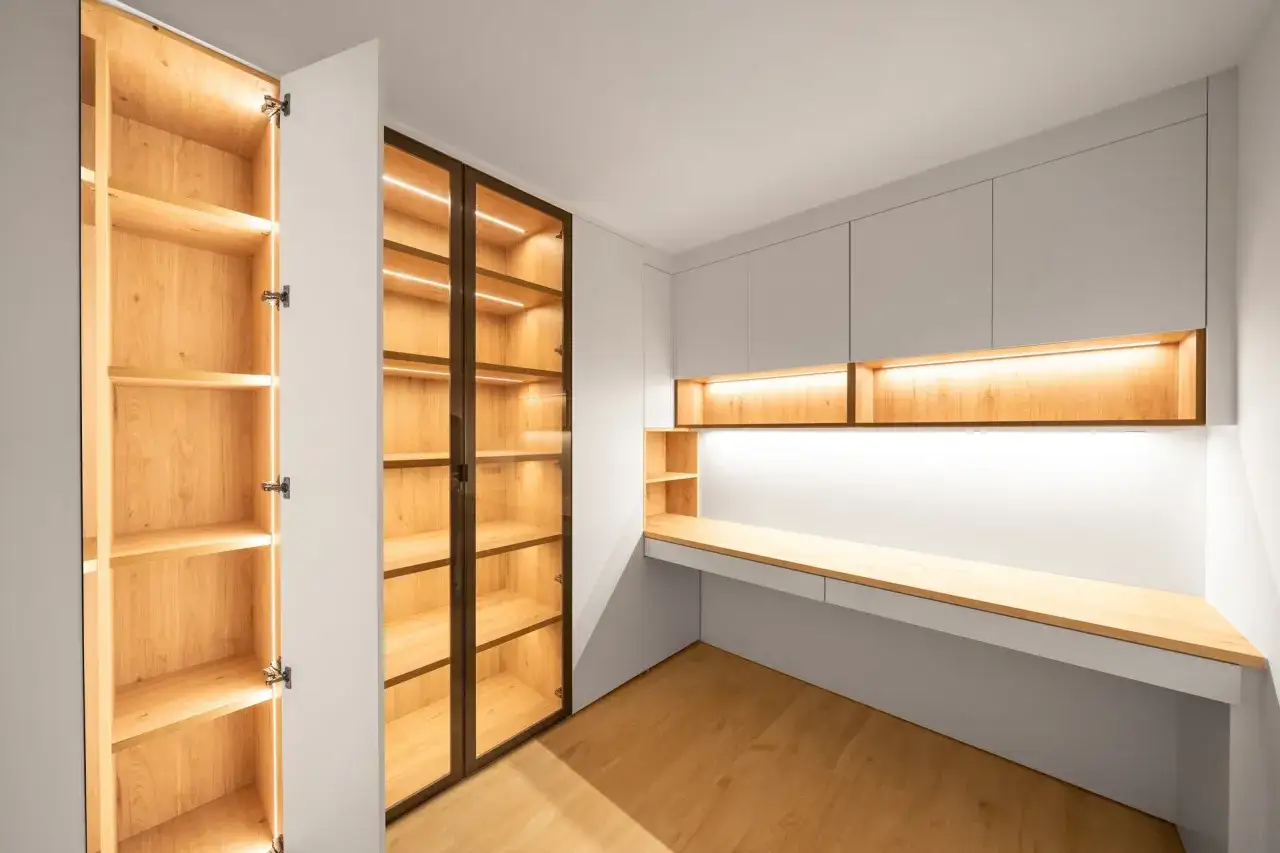 Armarios a medida con iluminación LED cálida, uno abierto de madera y otro acristalado, junto a un escritorio moderno.