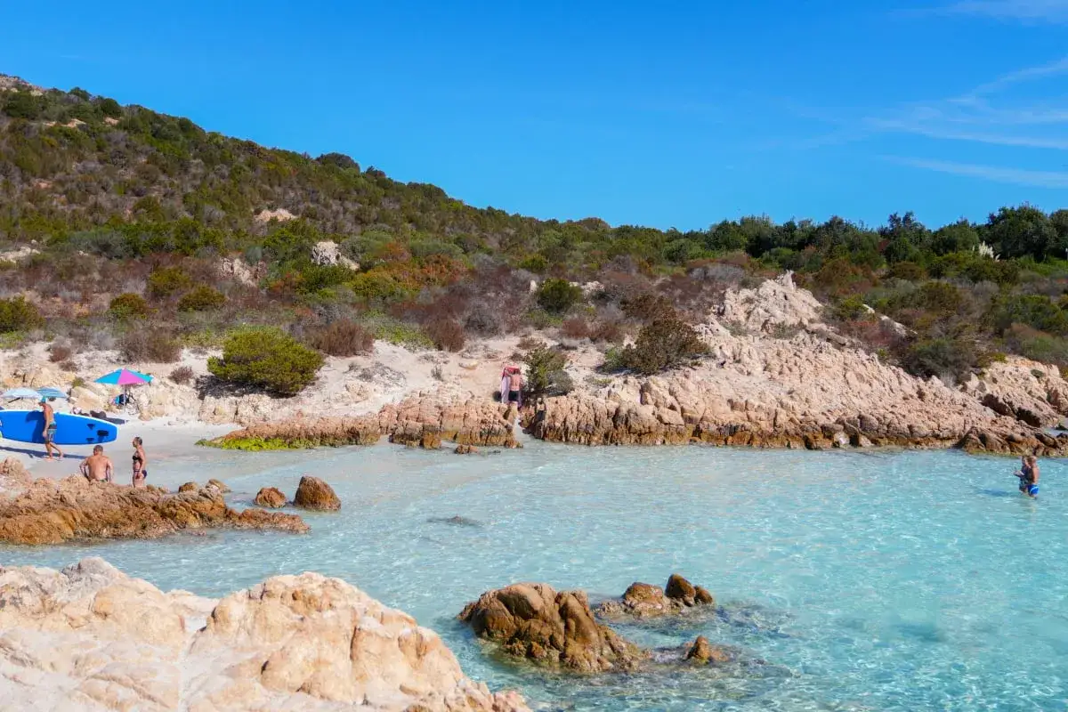 Sardinien im Oktober: Ein Mann mit Surfbrett am Strand, das Wetter ist sonnig und das Meer ist kristallklar.