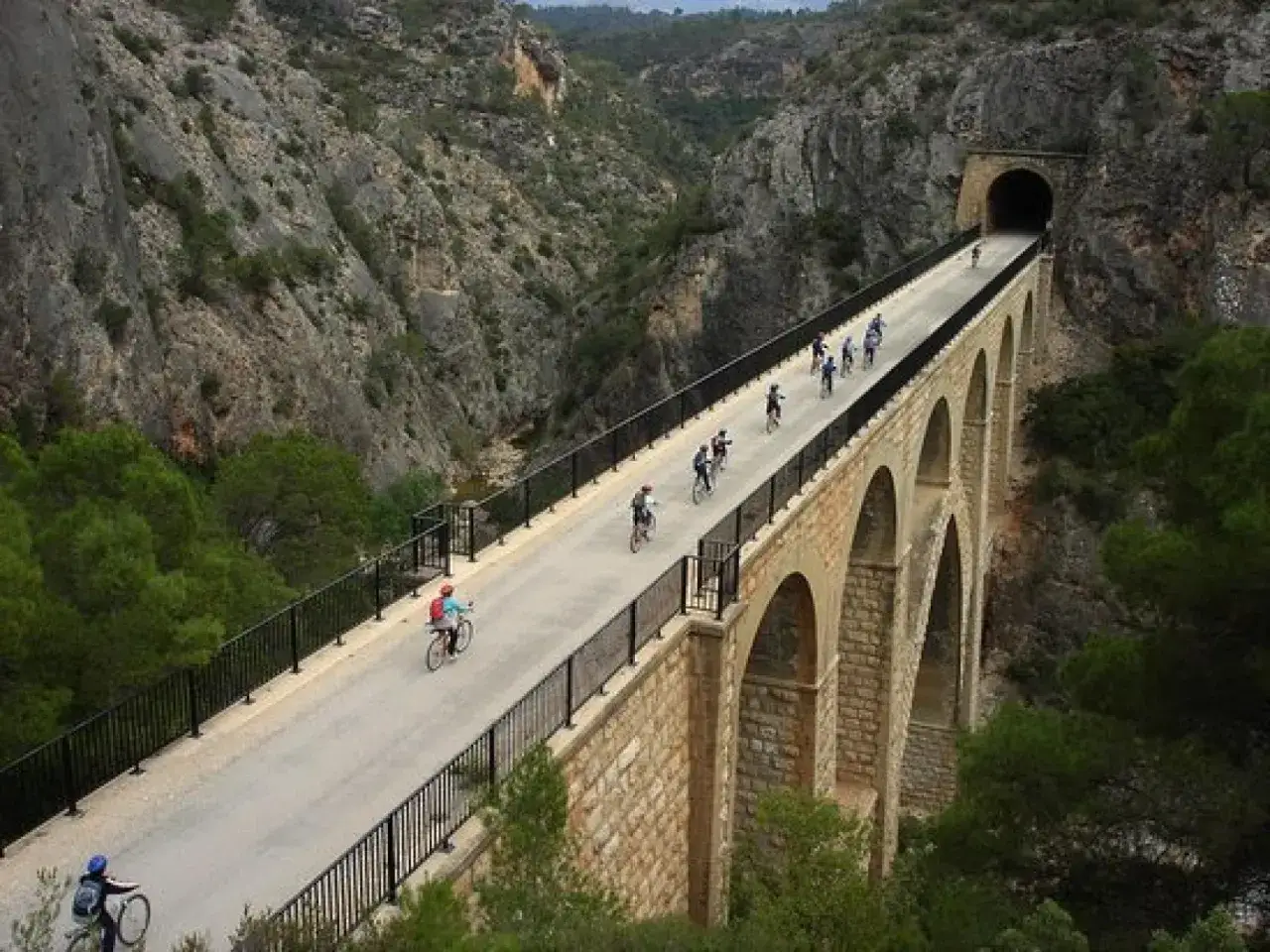 Ciclistas disfrutan de las **rutas ebike** por un puente antiguo con arcos, atravesando un cañón rocoso hacia un túnel.