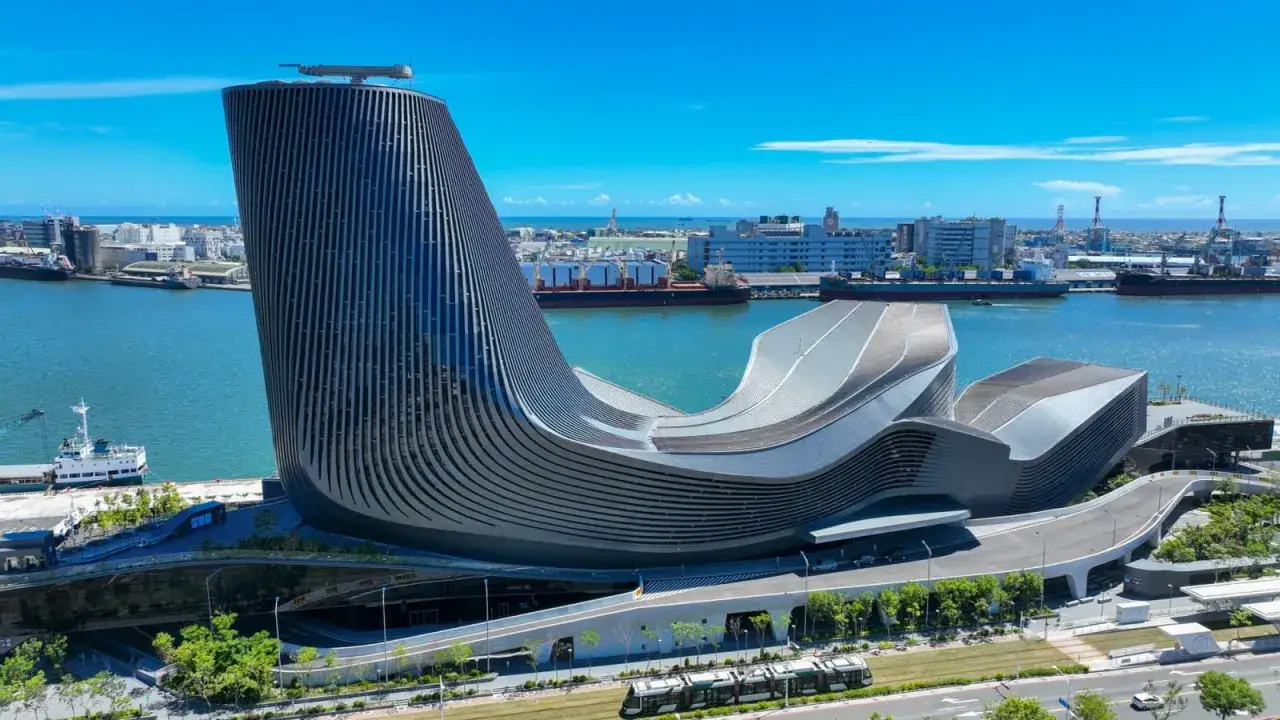 Moderne Architektur am Kaohsiung Port: Ein geschwungenes Gebäude mit vertikalen Lamellen ragt neben Frachtschiffen und dem blauen Meer auf.
