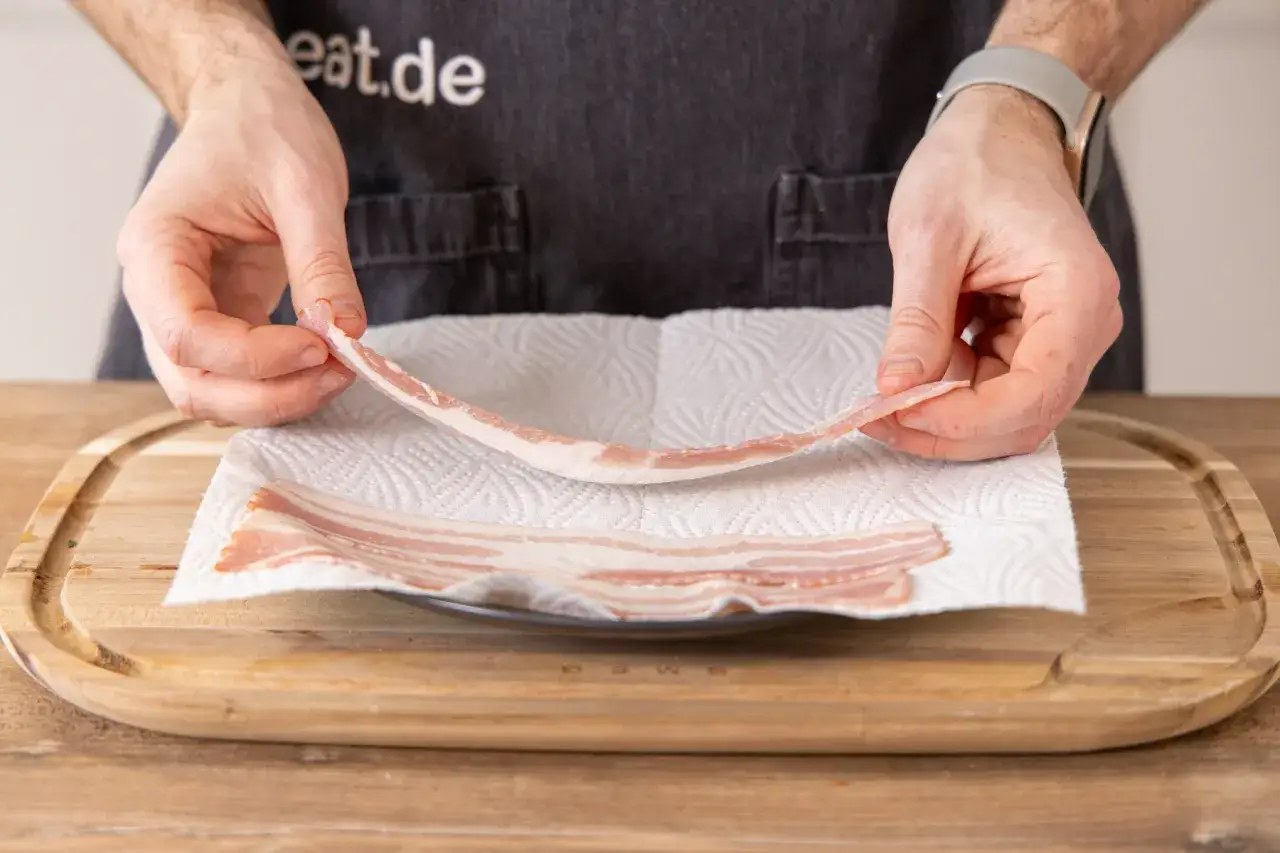 Hände legen rohen Bacon auf Küchenpapier, bereit für die Mikrowelle.