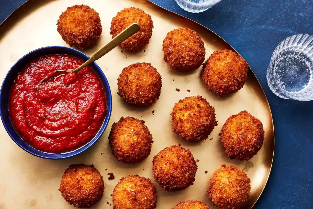 Arancini dor&eacute;s et croustillants dispos&eacute;s sur un plateau, accompagn&eacute;s d'une sauce pour arancini rouge vif dans un bol bleu.