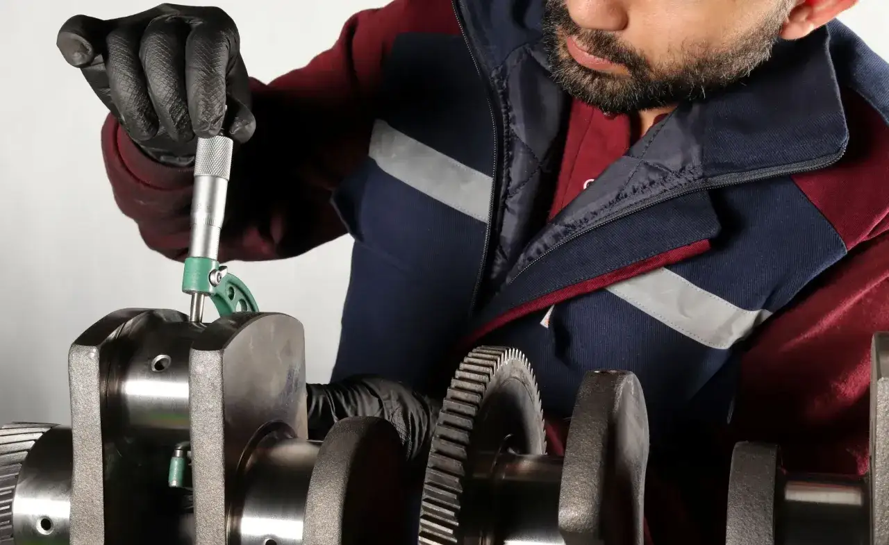 Mechanik w czarnych rękawiczkach precyzyjnie mierzy suwmiarką wał korbowy.