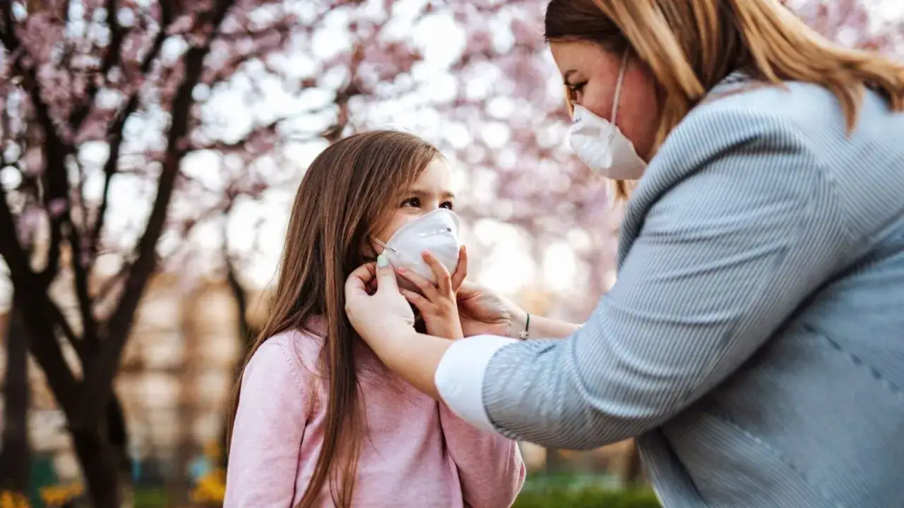 Madre ajusta mascarilla FFP2 a su hija bajo un árbol en flor. La mascarilla FFP2 sirve para proteger de partículas finas.