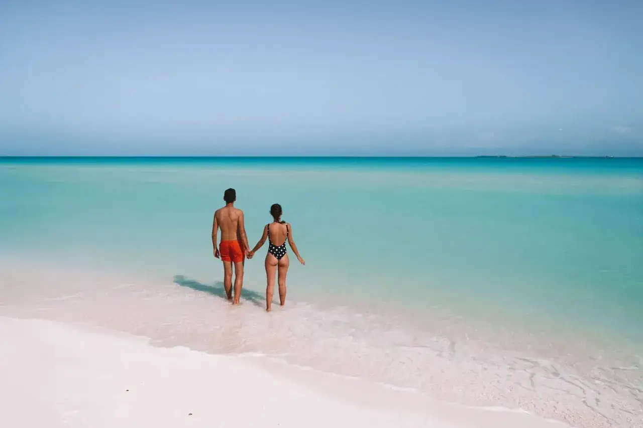 Pareja cogida de la mano camina hacia el mar turquesa en una playa de arena blanca. ¡El momento perfecto para saber cuándo viajar a Cuba!