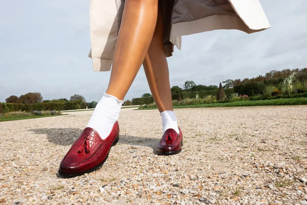 Pieds chaussés de mocassins rouges vernis, ornés de pompons, marchant sur un chemin de gravier. Le rouge vif de ces chaussures suggère audace et passion.