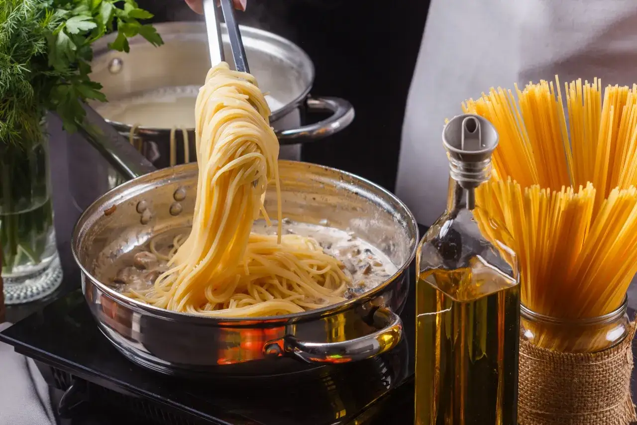 Szef kuchni dodaje do sosu grzybowego idealny makaron al dente. Obok butelka oliwy i pęczek spaghetti.