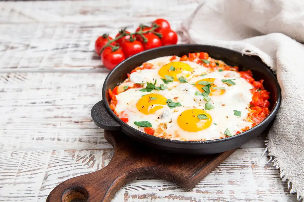Ein köstliches Shakshuka Rezept in einer Gusseisenpfanne, garniert mit Petersilie. Daneben ein Bund Kirschtomaten.