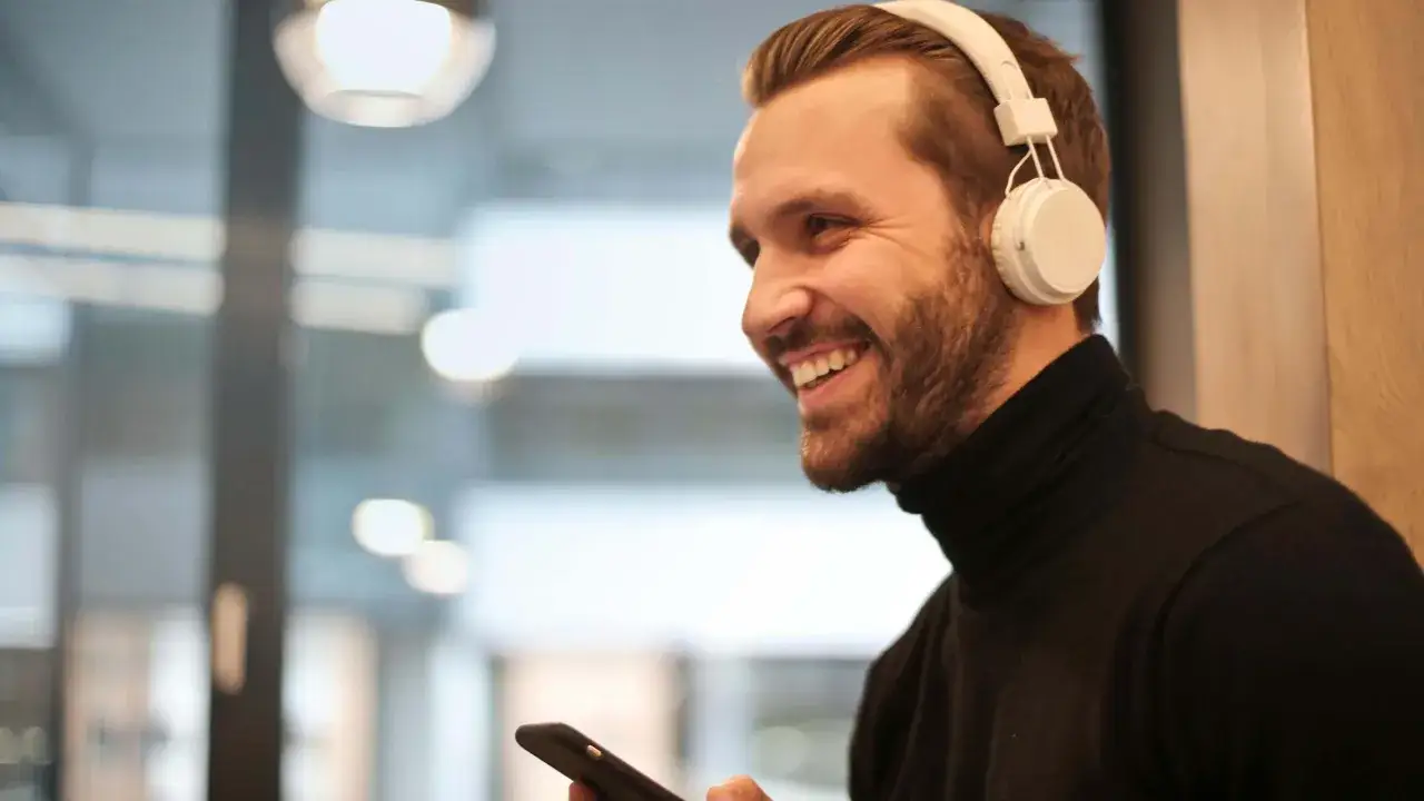 Uśmiechnięty mężczyzna w białych słuchawkach na uszach i czarnym golfie trzyma telefon. Dowiedz się, jak sparować słuchawki z urządzeniem.