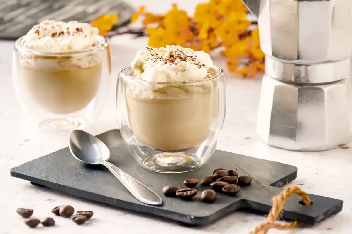 Delicioso mousse de caf&eacute; con crema batida y espolvoreado de cacao, servido en vasos de doble pared. Una cafetera italiana y granos de caf&eacute; completan la escena.